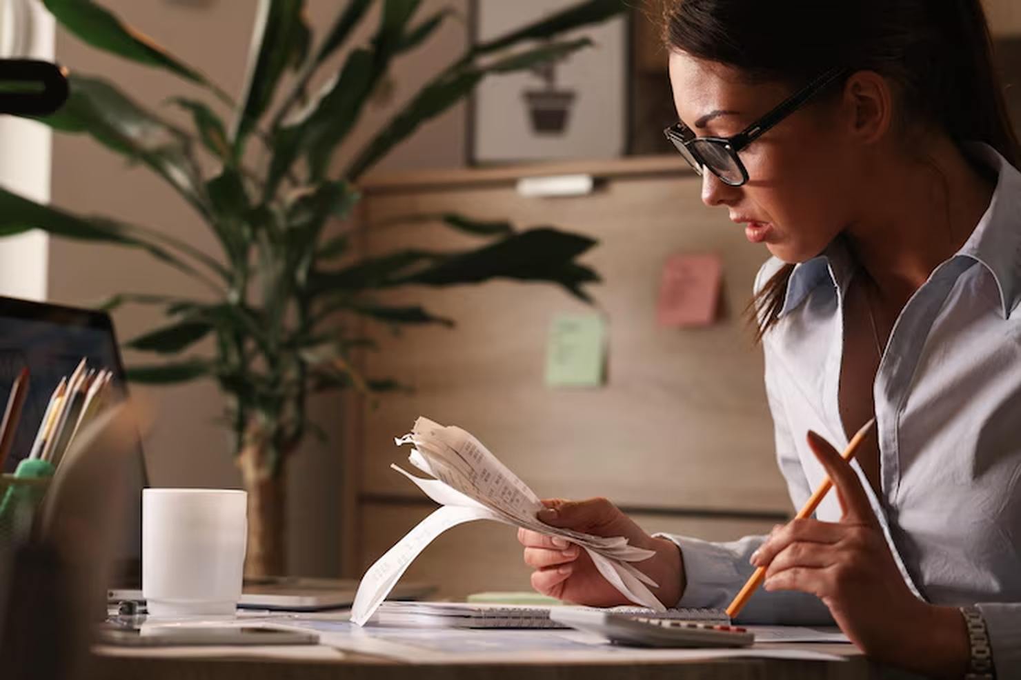 A woman wearing glasses sits at a desk reviewing receipts and using a calculator, likely organizing her finances or managing expenses related to personal credit, with a laptop and office supplies nearby.