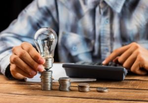 A person calculating energy bills while holding a light bulb over stacks of coins, symbolising cost savings and efficient electricity management in the UK.
