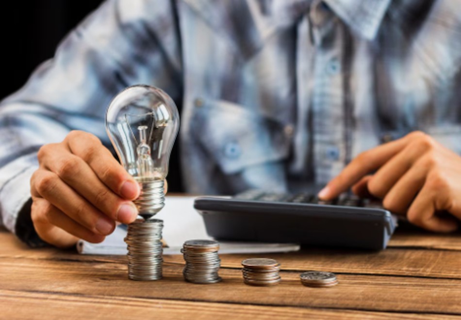 A person calculating energy bills while holding a light bulb over stacks of coins, symbolising cost savings and efficient electricity management in the UK.