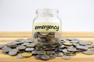 A glass jar labeled “emergency” filled with coins and surrounded by loose change, symbolizing saving money and building an emergency fund for financial security.