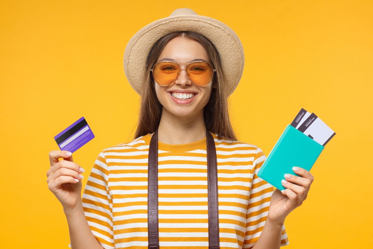 Smiling woman holding a credit card and travel tickets against a bright yellow background, representing the use of points programs and discounts for affordable UK travel.